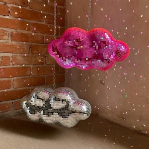 May include: Two cloud-shaped disco ball decorations hanging from chains. One is silver, the other is pink. The pink cloud has a gradient effect, transitioning from a darker pink to a lighter pink. The background is a brick wall and a beige wall.