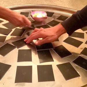 May include: A large, round, silver tray holds numerous black cards, arranged in a circular pattern. Hands reach towards the cards, suggesting a game or activity. A small candle holder with a lit candle sits in the center.