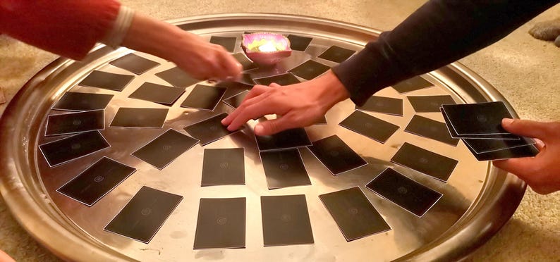 May include: A large, round, silver tray holds numerous black cards, arranged in a circular pattern. Hands reach towards the cards, suggesting a game or activity. A small candle holder with a lit candle sits in the center.
