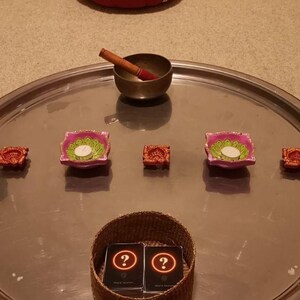 May include: A large circular silver tray holds a metallic singing bowl with a wooden striker, two pink lotus-shaped candle holders with white tea lights, two small orange decorative bowls, and a woven basket containing two black cards with question marks.