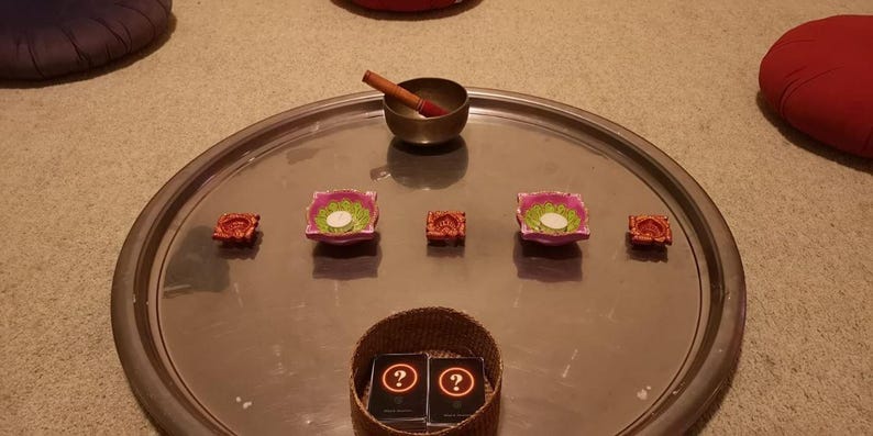 May include: A large circular silver tray holds a metallic singing bowl with a wooden striker, two pink lotus-shaped candle holders with white tea lights, two small orange decorative bowls, and a woven basket containing two black cards with question marks.