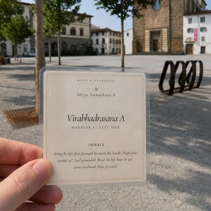 May include: A rectangular, cream-colored instructional card encased in a clear plastic sleeve. The text reads: BOND & AWARENESS, Surya Namaskara B, Virabhadrasana A, WARRIOR I LEFT SIDE, INHALE, bring the left foot forward between the hands.