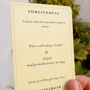 May include: A rectangular cream-colored card with rounded corners featuring black text. The top reads FORGIVENESS followed by the text I release only what my heart is ready to release. Below is the question What is still asking to be felt? and an inverted section reading REPRESSION with the text I bury what still longs to be heard. What am I protecting myself from feeling?