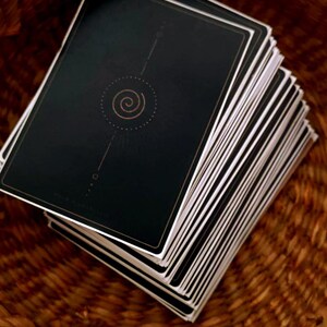 May include: A stack of tarot cards with black backs and white edges rests inside a woven basket. The card backs feature a gold spiral design and a thin gold border. The basket is made of brown woven material.
