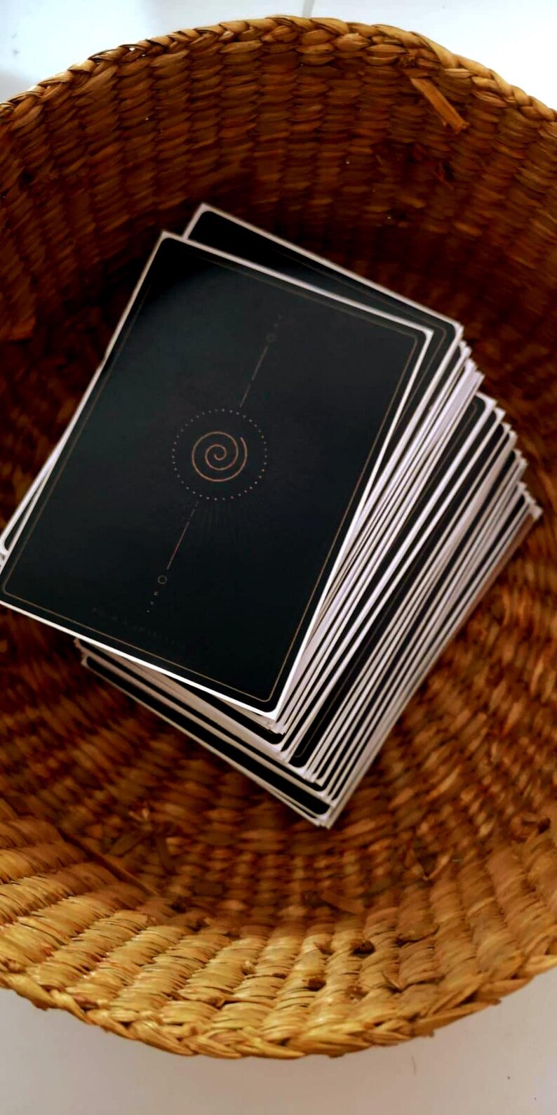 May include: A stack of tarot cards with black backs and white edges rests inside a woven basket. The card backs feature a gold spiral design and a thin gold border. The basket is made of brown woven material.