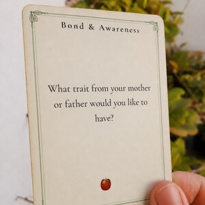 May include: A rectangular off-white card with a thin green border and decorative corners. The text reads Bond & Awareness followed by the question: What trait from your mother or father would you like to have? A small red apple icon is at the bottom.