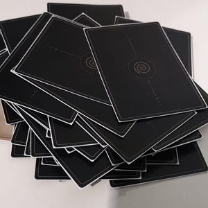 May include: A stack of black playing cards with a white border. The back of each card features a gold spiral design with radiating lines and small dots. The cards are arranged in a slightly fanned-out pile.