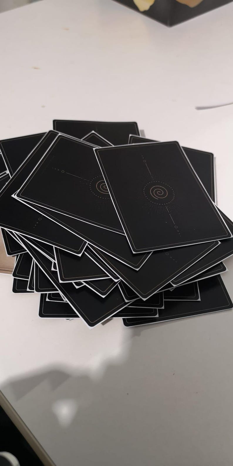 May include: A stack of black playing cards with a white border. The back of each card features a gold spiral design with radiating lines and small dots. The cards are arranged in a slightly fanned-out pile.