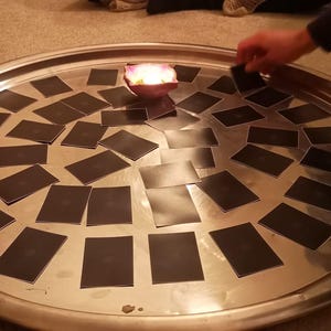May include: A large, round, silver tray with a candle in the center, surrounded by many black cards. Several hands are visible, interacting with the cards, suggesting a game or ritual. The scene is lit by the candle.
