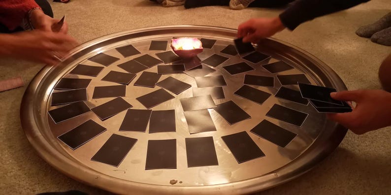 May include: A large, round, silver tray with a candle in the center, surrounded by many black cards. Several hands are visible, interacting with the cards, suggesting a game or ritual. The scene is lit by the candle.
