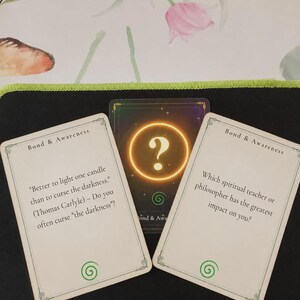 May include: Three rectangular cards from a Bond & Awareness deck. Two white cards feature black text and a green spiral icon. The left card quotes Thomas Carlyle, while the right asks about spiritual teachers. A central dark card displays a glowing gold question mark.