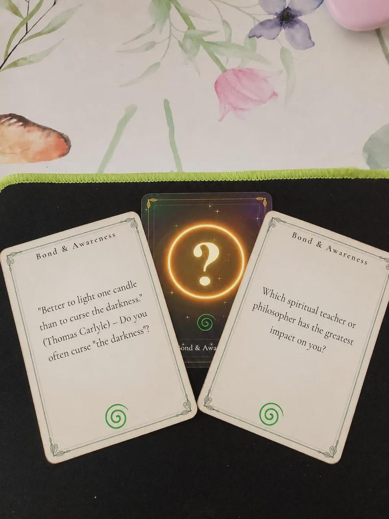 May include: Three rectangular cards from a Bond & Awareness deck. Two white cards feature black text and a green spiral icon. The left card quotes Thomas Carlyle, while the right asks about spiritual teachers. A central dark card displays a glowing gold question mark.