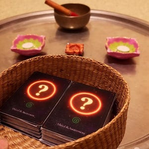 May include: A woven basket containing two stacks of black cards with a glowing orange question mark and the text Mind & Awareness. In the background, a metal tray holds a small bronze bowl with a wooden striker and two pink candle holders.