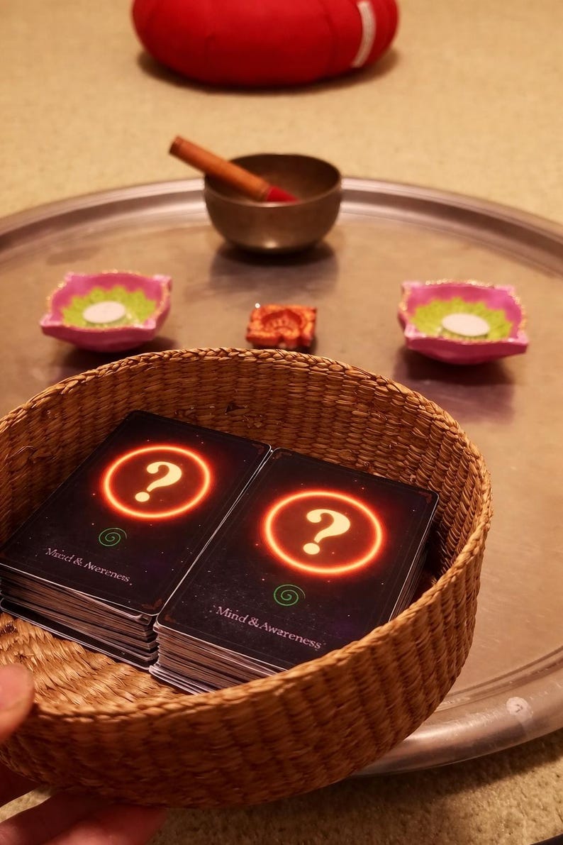 May include: A woven basket containing two stacks of black cards with a glowing orange question mark and the text Mind & Awareness. In the background, a metal tray holds a small bronze bowl with a wooden striker and two pink candle holders.