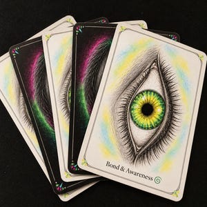 May include: A fan of rectangular oracle cards with white borders and rounded corners. The top card features a detailed illustration of a human eye with a vibrant green and yellow iris, surrounded by soft blue and yellow shading. The text Bond & Awareness is printed at the bottom.