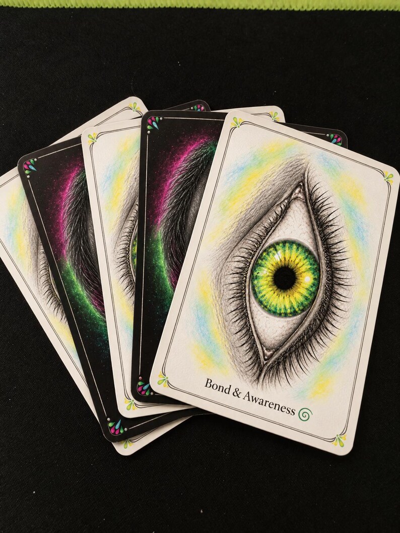 May include: A fan of rectangular oracle cards with white borders and rounded corners. The top card features a detailed illustration of a human eye with a vibrant green and yellow iris, surrounded by soft blue and yellow shading. The text Bond & Awareness is printed at the bottom.