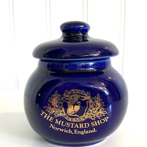 May include: A deep cobalt blue ceramic jar with a matching lid. The front features a gold-colored graphic of a bull head surrounded by a laurel wreath, with the text "THE MUSTARD SHOP Norwich, England." printed in gold lettering below the image.