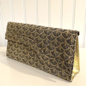 May include: A rectangular clutch bag featuring a metallic gold and black scalloped fan pattern across the exterior. The bag has a structured fold-over flap design with a shimmering finish and a solid gold interior lining visible on the side panel.