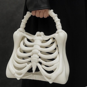 May include: A white handbag shaped like a human rib cage with a matching bone-inspired handle. The structure features moulded, curved ribs extending from a central sternum piece, creating a hollow, sculptural design in a matte off-white finish.