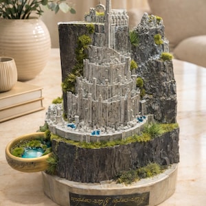 Puede incluir: Una maqueta detallada de Minas Tirith, una ciudad de El Señor de los Anillos, con una alta torre blanca y estructuras de piedra en capas. El modelo incluye una pequeña piscina de agua y está sobre una base rocosa con detalles verdes.