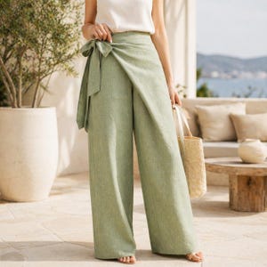Puede incluir: Pantalones palazzo verde claro con cinturón a juego. Los pantalones de pernera ancha están hechos de una tela texturizada y se combinan con una parte superior blanca. Se sostiene un bolso de mano de paja. Los pantalones son de un color verde suave y apagado.