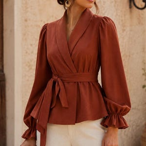 Op de afbeelding: Een roestkleurige wikkelblouse met lange mouwen met ruches aan de manchetten, een sjaalkraag en een bijpassende stoffen ceintuur in de taille. Het kledingstuk wordt gecombineerd met een effen witte broek voor een gestructureerd en samenhangend geheel.