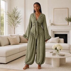 May include: An olive green two-piece outfit. The top has long sleeves with ruffled cuffs and a tie closure. The pants are wide-legged with a gathered waist and drawstring ankles. The outfit is made of a textured fabric and is worn with tan sandals.