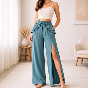May include: A woman's outfit comprising a cream-coloured crop top and teal wide-leg trousers with a high slit. The trousers have a ruffled waistline with a bow. The outfit is paired with sandals.