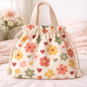 Puede incluir: Una bolsa con cordón de color crema con un estampado floral y de corazones. La bolsa presenta un diseño repetido de flores coloridas en tonos naranja, amarillo y verde, junto con corazones rojos. Tiene un asa a juego y cierre de cordón.