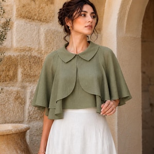 May include: An olive green capelet featuring a rounded collar and a layered, flared silhouette. The garment has a smooth texture and falls to the waist, paired with a plain white skirt. The design includes a front opening and short, wide sleeves.