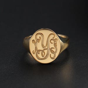 May include: A gold signet ring featuring a polished band and a circular face. The center of the ring is engraved with a textured, ornate monogram consisting of the letters X, Y, and Z in a decorative, interconnected script style.