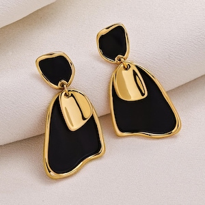 May include: A pair of gold-toned earrings with a black enamel inlay. Each earring features a small, rectangular top piece and a larger, abstract-shaped bottom piece. The earrings have a polished finish and a modern design.