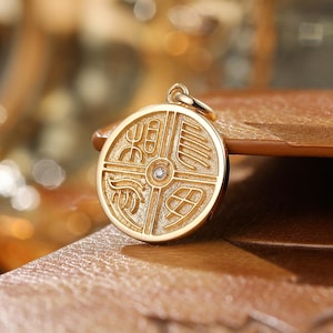May include: A circular gold pendant featuring a textured surface divided into four quadrants by raised lines. Each quadrant contains a distinct engraved symbol, and a small, clear, round gemstone is set in the center of the pendant.