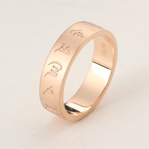 Könnte beinhalten: Ein Ring aus Roségold mit einer Reihe eingravierter Symbole auf der Außenfläche. Die Innenseite des Rings ist deutlich mit dem Text Au750 gekennzeichnet, was den Reinheitsgrad des Metalls angibt. Der Ring hat eine glatte, polierte Oberfläche.