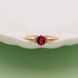 May include: A rose gold ring featuring a central oval-cut deep red gemstone flanked by two small clear stones on each side. The band has a delicate beaded texture that runs along the entire circumference, creating a refined and detailed appearance.