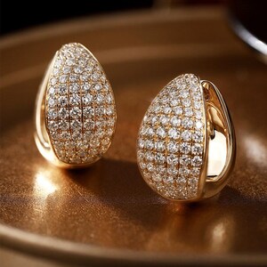 Pode incluir: Um par de brincos dourados em forma de gota. Cada brinco é incrustado com numerosas pedras preciosas pequenas e transparentes. Estão sobre uma superfície dourada e brilhante.