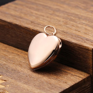 May include: A polished rose gold heart-shaped locket pendant with a smooth, reflective surface. The locket features a small, circular jump ring at the top for attachment to a chain, showcasing a simple, clean design with no visible engravings.