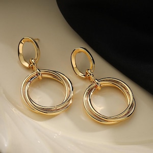 May include: A pair of gold-coloured earrings. Each earring features a small, oval-shaped top connected to a series of intertwined gold rings. The earrings have a polished, reflective surface, and are displayed on a white surface.