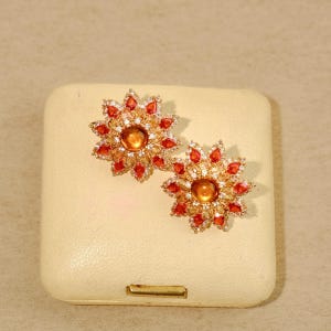 Pode incluir: Um par de brincos dourados. Cada brinco apresenta uma pedra central laranja-castanha, rodeada por pequenas pedras brancas. O anel exterior é composto por pedras em forma de pétala vermelho-laranja, criando um design floral.