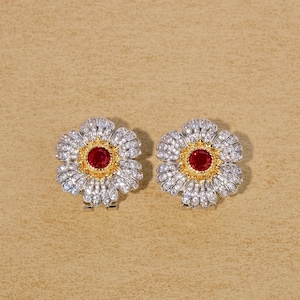 Op de afbeelding: Een paar bloemvormige oorbellen. Elke oorbel heeft een centrale rode edelsteen omgeven door een goudkleurige ring. De bloemblaadjes zijn bezet met kleine, heldere edelstenen. De oorbellen staan tegen een neutrale achtergrond.