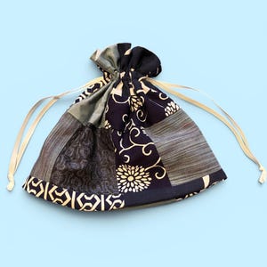 May include: A drawstring pouch crafted from a patchwork of fabric panels in navy blue, grey, and white. The textile features a mix of patterns including floral motifs, geometric shapes, and fine linear textures, finished with thin cream-colored cords.