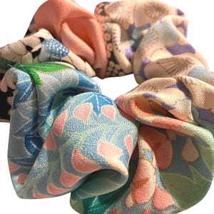 以下が含まれることがあります： ピンク、ブルー、グリーン、クリーム色を基調とした、様々な花柄が特徴的なカラフルなシュシュ。生地はシルクまたは同様の軽量素材で、柔らかくギャザーの入った質感です。このヘアアクセサリーは、ポニーテールやお団子ヘアをまとめるためにデザインされています。