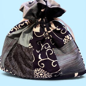 May include: A drawstring pouch constructed from a patchwork of fabrics in navy blue, light blue, and grey. The bag features a repeating white floral pattern on the navy sections, solid grey panels, and a textured grey fabric, closed with a white ribbon.