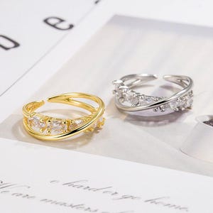 Puede incluir: Dos anillos abiertos, uno dorado y otro plateado, cada uno adornado con piedras preciosas claras y brillantes. El anillo dorado presenta un diseño de doble banda, mientras que el anillo plateado tiene un estilo entrecruzado. Ambos anillos están sobre un fondo suave y neutro.