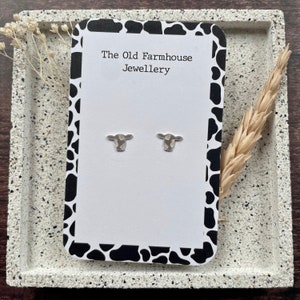 May include: A white card with black and white cow print border. The card says "The Old Farmhouse Jewellery" and features two silver cow head stud earrings.