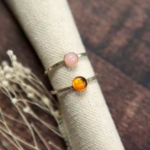 May include: Two silver rings with round gemstones. The top ring has a light pink gemstone and the bottom ring has an amber gemstone.