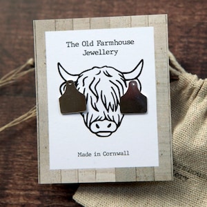 May include: A pair of silver earrings shaped like cow tags, displayed on a card with a black and white illustration of a Highland cow. The card reads "The Old Farmhouse Jewellery" and "Made in Cornwall".