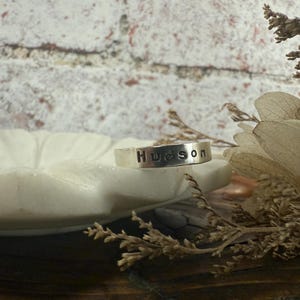 May include: A silver ring with the name "Hudson" stamped across the top. The ring is displayed on a white, petal-shaped dish, with dried floral accents. The background features a textured, white brick wall.