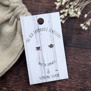 May include: Silver stud earrings with a teacup and teapot design. The earrings are presented on a white card with the text "THE OLD FARMHOUSE JEWELLERY" and "MADE IN CORNWALL". A beige drawstring bag is in the background.