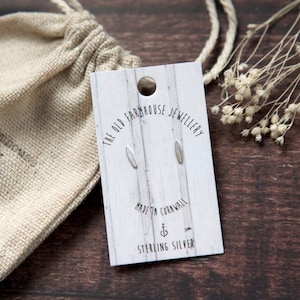May include: A pair of sterling silver earrings in the shape of a small, thin oval. The earrings are displayed on a white card with the text "The Old Farmhouse Jewellery" and "Made in Cornwall".